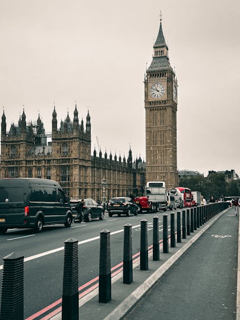 A street scene in London showing the historic Big Ben clock tower and the Houses of Parliament in the background with a cloudy sky. In the foreground, a traffic lane contains various vehicles includin